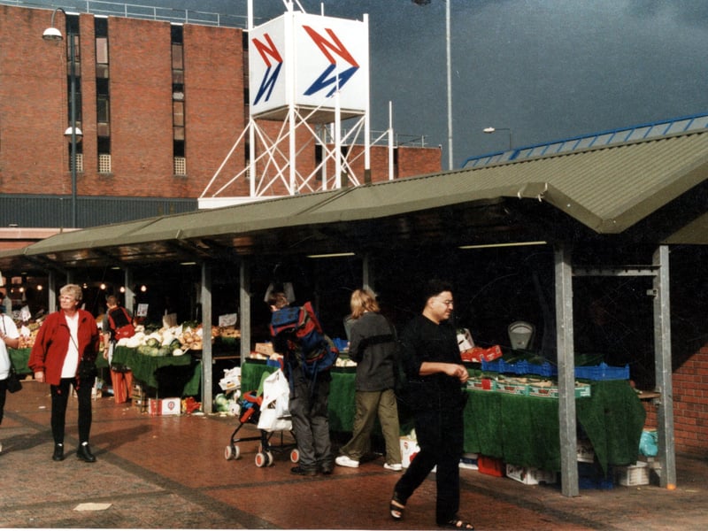This view of the outdoor market shows a row of fruit and vegetable stalls. In the background is the Leeds City bus station with the National Express sign prominent. In the distance is Millgarth Police Station. Pictured in September 1999.