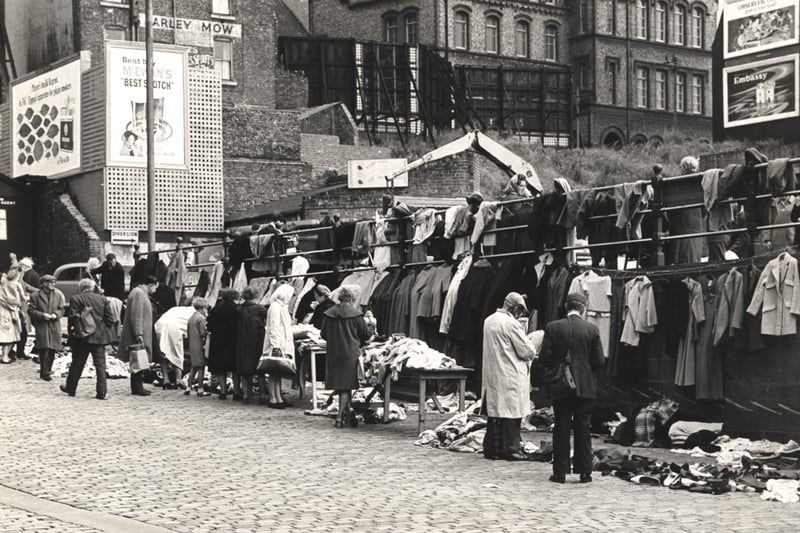 A view of Paddy's Market taken in 1965. The photograph shows clothes for sale hanging from the railings on tables and on the ground. Vendors and customers are standing beside the clothes. The rear of the Barley Mow pub and a school can be seen in the background.