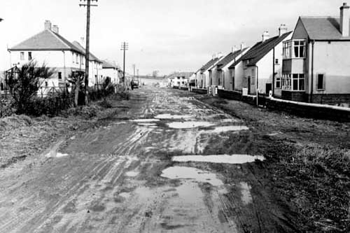 A view looking north along Kingsley Avenue in February 1953. Very muddy road with houses and wooden gates on either side. People, telegraph poles and a truck can be seen