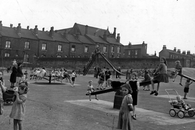 The recreation ground on Beckett Street in June 1949. Houses line the horizon. A slide, swings, roundabout and see-saws are visible. A large number of children are playing. Adults can be seen supervising. A pram is visible on the right with two children in it.