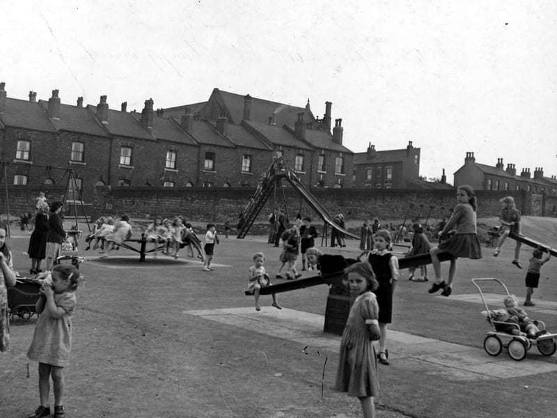 The recreation ground on Beckett Street in June 1949. Houses line the horizon. A slide, swings, roundabout and see-saws are visible. A large number of children are playing. Adults can be seen supervising. A pram is visible on the right with two children in it.