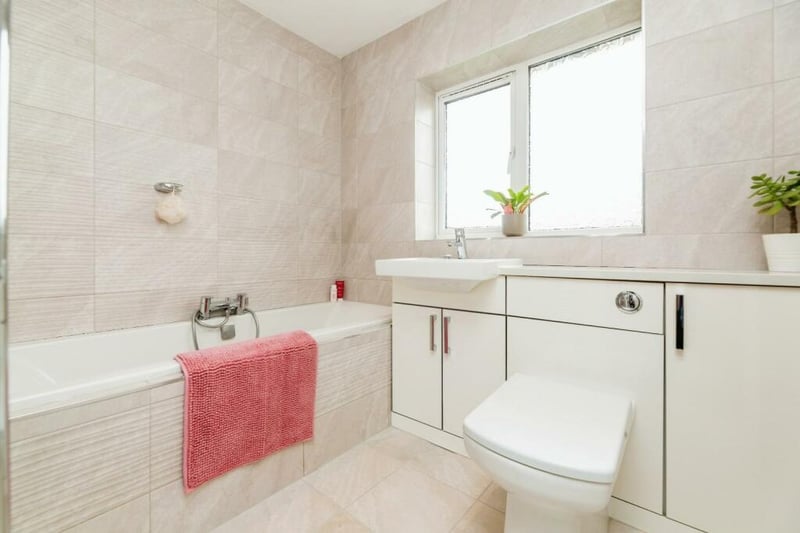A serene family bathroom hosting natural stone textured wall tiles and cream stone tiled flooring, white gloss vanity unit with inset ceramic sink and low flush WC, bath with chrome telephone mixer tap, wall mounted chrome heated towel rail, inset spotlights, extractor fan and frosted uPVC window.