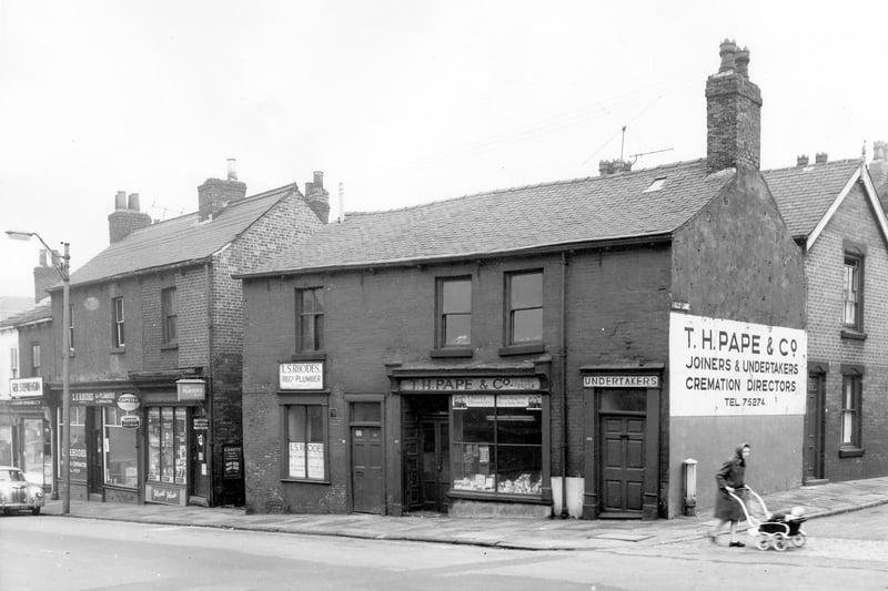Shops on Beeston Hill in Octobeer 1963. They include L.S. Rhodes registered plumbers and contractors on the left. On the right is A & T Hudson grocers. There is an alleyway between buildings that leads to Una Mount. On the right is Folly Lane where a woman in a headscarf and overcoat pushes a child's pram across the street. This section of Folly Lane where it meets Beeston Road was later demolished making Folly Lane accessable via Lady Pit Lane on the other side.