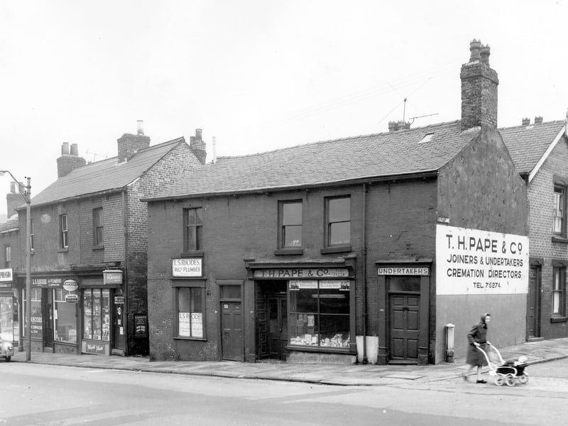 Shops on Beeston Hill in Octobeer 1963. They include L.S. Rhodes registered plumbers and contractors on the left. On the right is A & T Hudson grocers. There is an alleyway between buildings that leads to Una Mount. On the right is Folly Lane where a woman in a headscarf and overcoat pushes a child's pram across the street. This section of Folly Lane where it meets Beeston Road was later demolished making Folly Lane accessable via Lady Pit Lane on the other side.