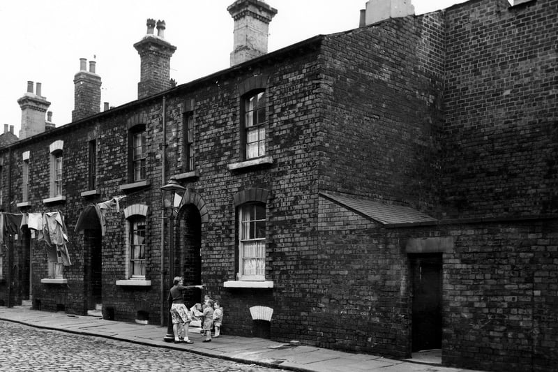 A row of back-to-back terraced houses with cellars on Arkwright Street in October 1963. On the right is a shared outside toilet block. On the left clothes hang on a line stretched across the street. In the centre a girl stands by a lamppost with three small children in short trousers.