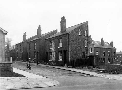 Raby Mount, seen from Sheepscar Street. Just seen on the left edge is the wall of a new Police Section House. At the end of Raby Mount, premises belonging to G.W. Binns, motor garages are visible. Raby Mount at this point comprised 3 pairs of houses. On the right is Yeomanry Street. Pictured in May 1958