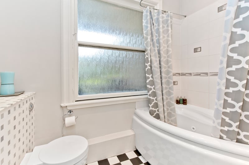 The principal bedroom’s en-suite is neutrally presented and finished with white tiles, framed by a border. It has a hidden-cistern toilet, a chic washbasin, downlit mirrored cabinet storage, and a whirlpool corner bath with overhead shower.