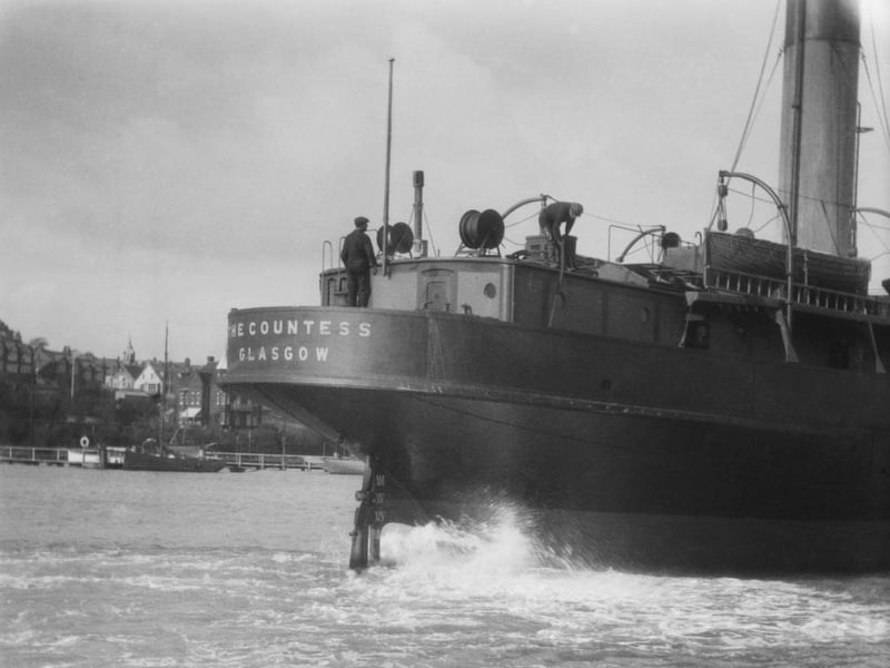 Two crewmen at work onboard the steamship 'The Countess Glasgow' as it powers through the water passing a harbour and town, English coast, circa 1924. 