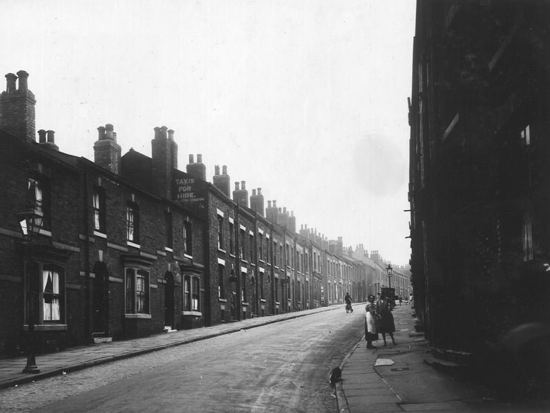Portland Crescent, showing length of street. Some houses have a bay window downstairs. Where gable of house on left is visible, a sign for Taxi Hire has been painted 'H Parish'. Two ladies on right face camera, man on bicycle in centre. The cobbled road has been partially covered in tarmac. Area now of Civic Hall building. Pictured in July 1930.