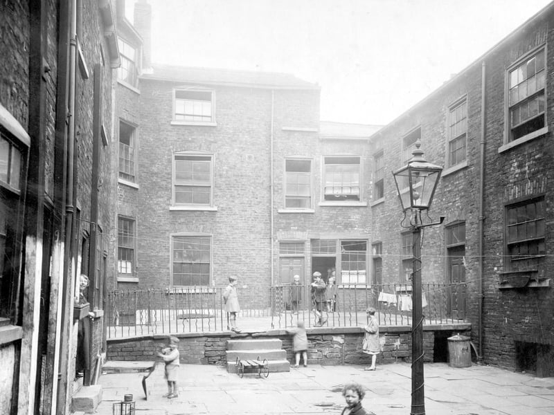 Hirst Square showing buildings on three sides. Several children are playing or standing in the square which is on two levels with steps and railings separating the two. Washing lines criss-cross the square and are tied to a street lamp in the middle. Pictured in July 1930.