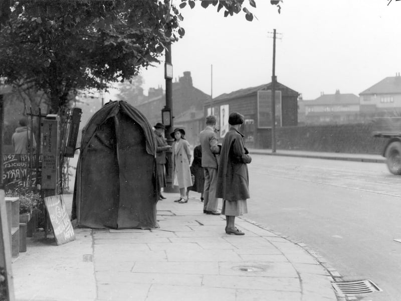 Tram stop on Otley Road at the corner with Shaw Lane, with people waiting in a queue. There is a workmen's shelter behind the queue. A vending machine for Wrigley's is on the railings. Semi-detached houses in view in the background.