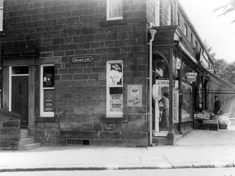 The corner of Shaw Lane and Otley Road, showing the side of no 54 Otley Road which is chemist shop of F C Stock with advertising signs and a weighing machine outside. Also shown is the outside display of J T Baxendale greengrocer, with a shop assistant. Pictured in August 1939.