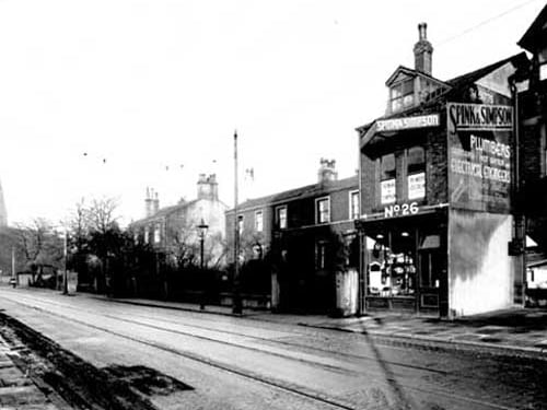 Premises of Spink and Simpson plumbers and electricians are at number 30 (number 26 on photo is not property number). To left number 32, residence and office of Joseph Leonard Iredale surgeon. Number 34 William Cuthbert Morton, surgeon. Houses set back from road with gardens. On left spire of Headingley Hill Congregational Church. Road is part cobbles, with tramlines. Pictured in November 1937.