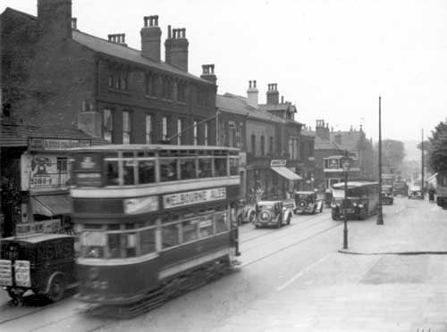 Stretch of Otley Road, looking towards North Lane. On the left, with telephone sign on wall is the fish shop of William Addison, this is number 15. Further to the right, shop with blind down is at number 31, Groocock and Son, grocers. The road is very busy, trams, a single decker bus, van and cars can be seen. Pictured in June 1937.