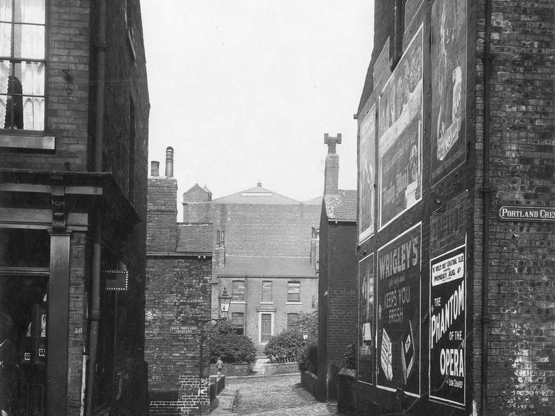 50 Portland Crescent on the left, looking through Back Portland Crescent. The house facing is on St James' Square. The streets are cobbled with streetlamps. Advertisements are on the end wall including one for "Phantom of the opera" and Wrigleys chewing gum. The area is now the site for Civic Hall. Pictured in July 1930.