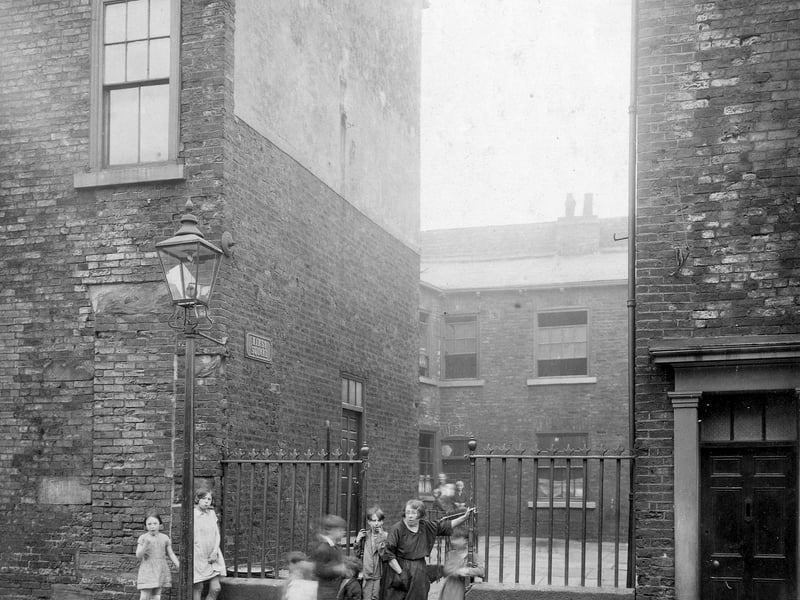 Hirst's Square entrance, off St. James Street. It has iron railings on a low wall between the adjacent buildings. In the entrance are a woman and several children, all poorly dressed. There is a street lamp by Hirst's Square. Site of the Civic Hall. Pictured in July 1930.