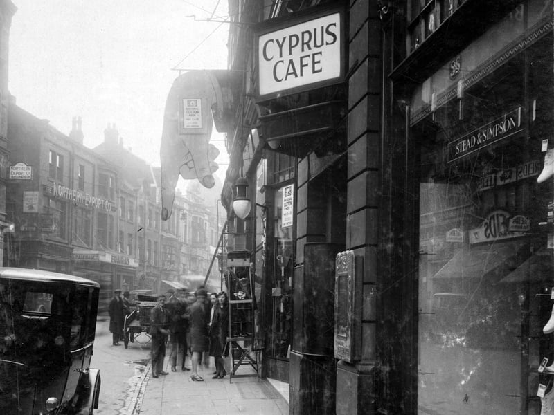 Kirkgate looking east. Stead and Simpson shoe shop on the corner of Briggate with a car parked on roadside. A sign for the Cyprus Cafe can be seen. This is 136 Kirkgate. M.C.Hitchens and Sons drapers was at 130-135 Kirkgate. A sign reading National Cotton Week May 5-10 can be seen on the board shaped like a hand pointing downwards. Pictured in May 1930.