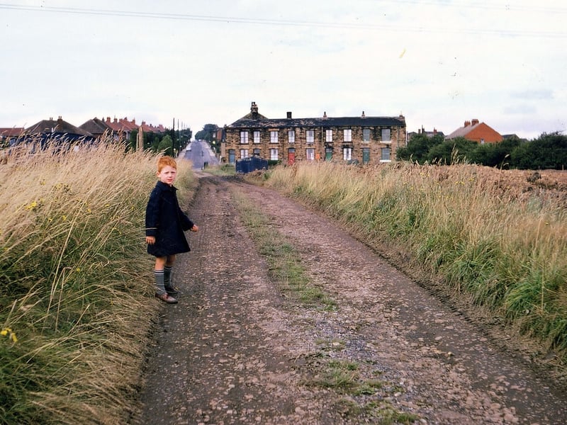 The view looks towards the junction of Haigh Moor Road with Batley Road. Haigh Moor Road is seen disappearing off into the distance and Batley Road cuts across in front of the stone built terraced properties. The little boy is standing on a continuation of haigh Moor Road as a cart track onto the fields of Haigh Hall Farm. Pictured in August 1967.
