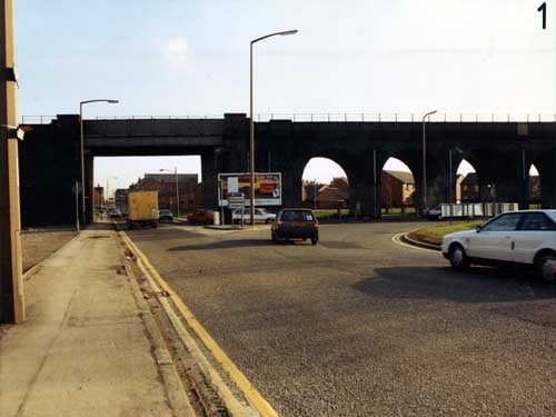 View looking south-east at the railway viaduct crossing Domestic Street, showing one rectangular archway and four round ones. An advertising hoarding in the middle displays a cigarette advertisement. Houses on Domestic Street and Ingram Gardens can be seen through the arches. Pictured in February 1991.