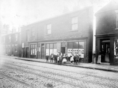 A parade of shops along the south side of Elland Road. 21 Elland Road in the centre of the photograph is R Wormald - hairdressers. Then there is a small alleyway into West Place and then 23 Elland Road is William Benn - draper. A large group of children are gathered in the centre.