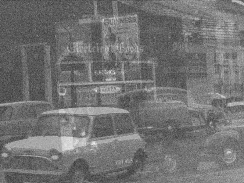 Flooding on Bondgate during the summer of 1961. A Morris Mini car makes its way through the water at the near end of the road. On the far side an electrical goods shop is seen on the left, then further back from the road is the Woolpack public house. Moving right the side wall of Garbutt and Mawson Ltd., ironmongers, is covered with advertisements including one for Guinness. On the right edge is the Bowling Green pub.