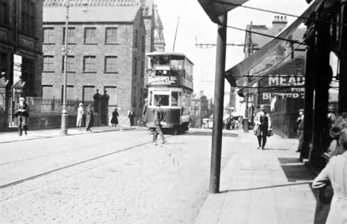 Queen Street taken from the front of Sam Clegg's shop looking north towards Morley Town Hall in July 1934. On the left hand side is the corner of the Queen Street Wesleyan Sunday School building with its iron railings and gateways. Across the road (Wesley Street) can be seen two buildings of Wesley Street Mill. The second of the two looks as if it is undergoing demolition as the roof is being stripped off.