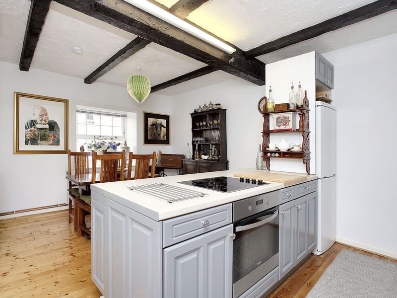 On the opposite side of the hall, the dual-aspect dining kitchen has similar proportions, pairing a white backdrop with wooden floorboards. It has a farmhouse-inspired design, and incorporates room for a table and chairs, alongside plenty of work surface space and ample cabinet storage. It also comes with integrated and freestanding appliances.