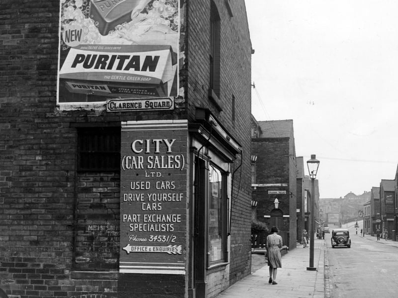 A view looking north along Sheepscar Street North past junctions with Clarence Square and Stanhope Square. Advertisements for Puritan soap and City Car Sales Ltd. People, cars and streetlamps are visible. A pub can be seen in the background. Pictured in September 1953.