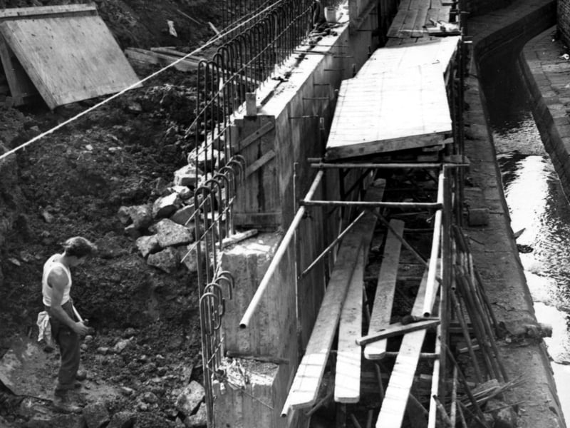 Rebuilding of a retaining wall at the G.P.O. garage. The beck can be seen on the right in a stone channel. The metal supports and concrete blocks of the wall are visible, together with scaffolding and timber walkways. Three workmen are visible. Pictured in September 1955.