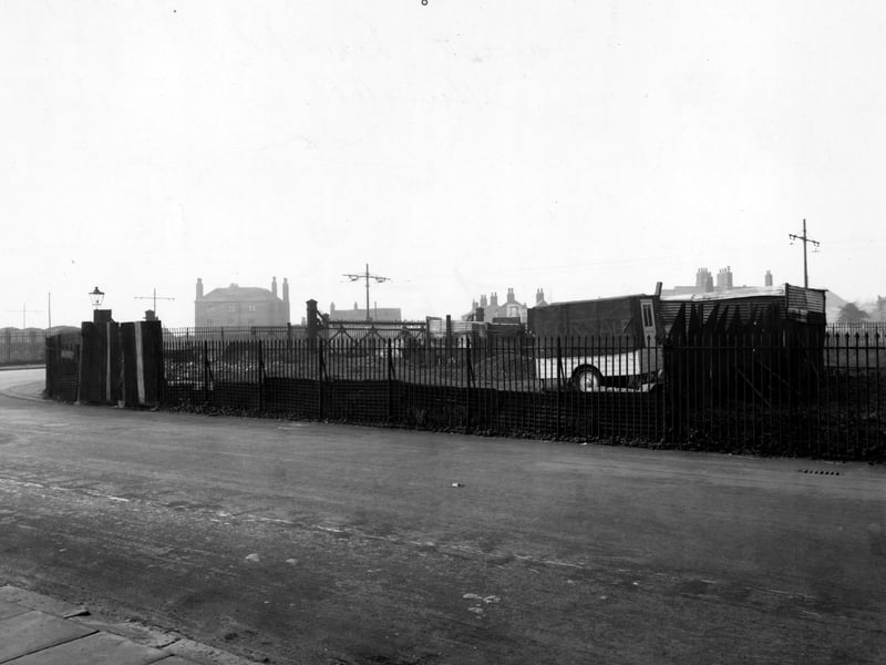 Moor Road in October 1947. This view shows J.E. Fawcett, Builders and Contractors. In the distance a few houses are visible. Around the site is a black cast iron fence, behind which is a trailer with a 'for sale' sign painted on it. Power lines run through the back of the site.