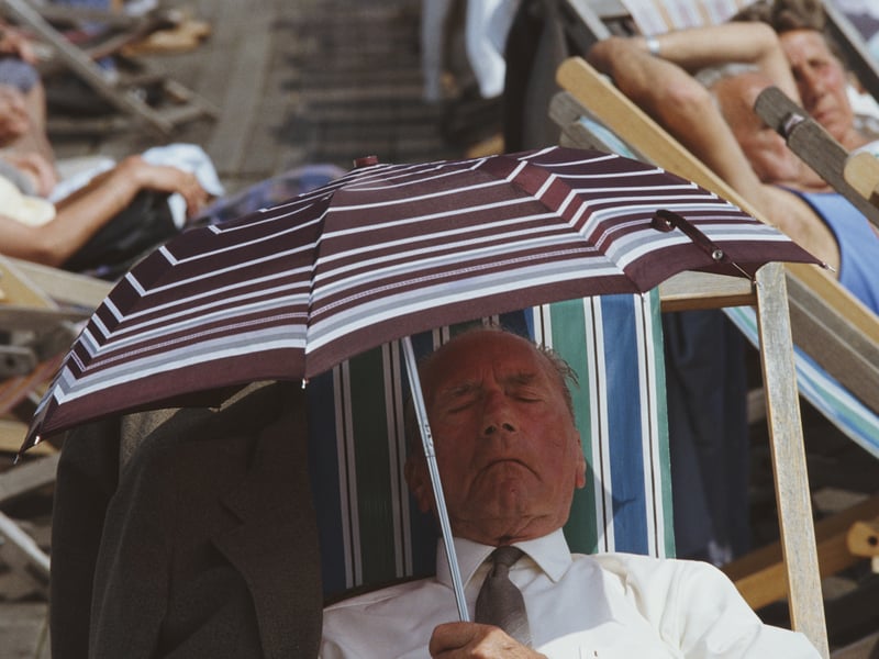 A man, wearing a shirt and tie, uses an umbrella to shelter from the sun whilst sitting in a deckchair amongst the crowds of people enjoying the warm weather on Blackpool's main beach
