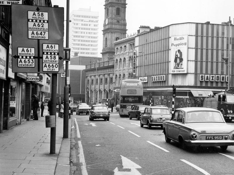 Duncan Street looking west towards Boar Lane, taken during the mid 1970s. On the left is the sign for the Whip public house in Hirst's Yard, the entrance to which is just off the picture. The first shop along is Modern Hairdressing for Men, with HMV Records and Jackson's Tailors further on. Shops on the right include Saxone shoes (advertising Hush Puppies), Modelia ladiewswear and Richard Shops ladieswear, followed by Holy Trinity Church.