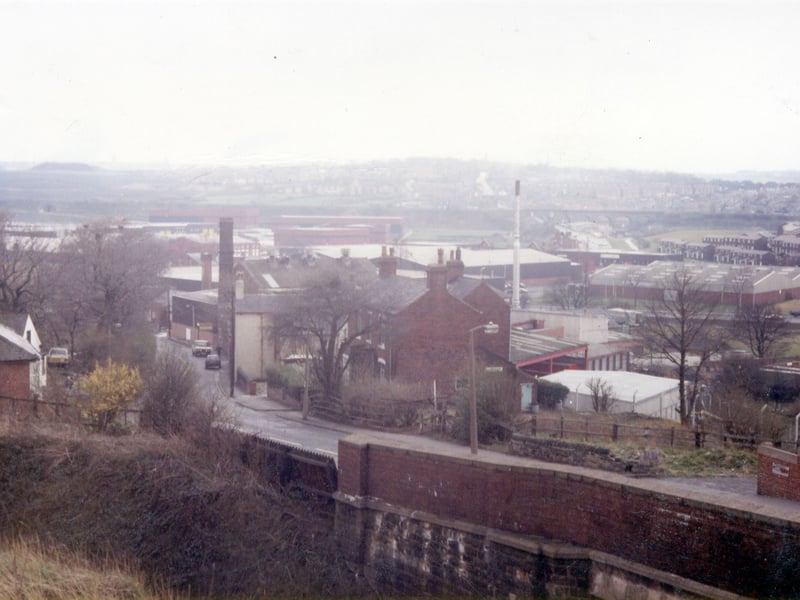Millshaw looking south-west with the railway bridge in the foreground on the right. Old buildings, including Holly House in the centre, are situated on the roadside while further back are newer industrial buildings. Cottingley Housing Estate is seen in the background on the far right with Churwell Viaduct behind and Churwell beyond this.