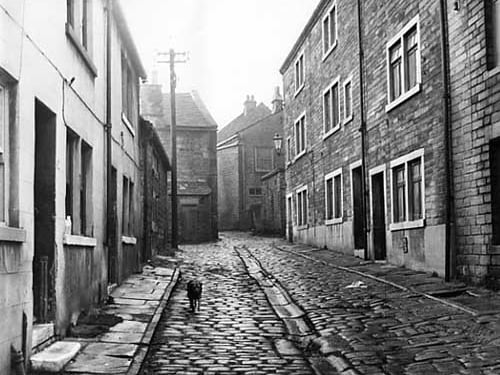 This photo shows one of the many yards and courts off Bramley Town Street. This is Wilson Place, presumably taking its name from an early inhabitant. Numbers 14 and 12 are visible on the left edge, both with cement rendered frontages. The yard is paved with setts and has an open gutter running down the centre. Pictured in January 1964.
