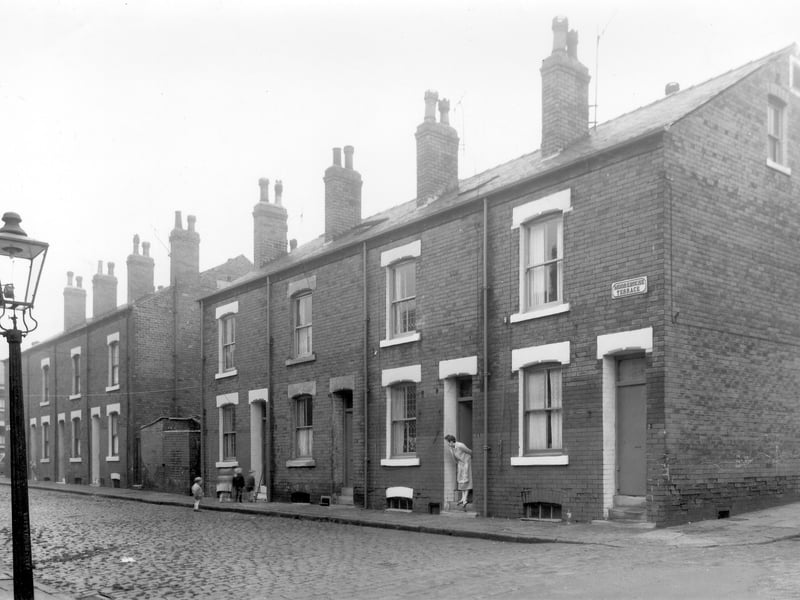 Back to back terraced housing on Moorehouse Terrace in October 1958. A lady looks out of the doorway of number 4 and children play outside number 8. The one storey brick buildings are outside toilet facilities for the street.