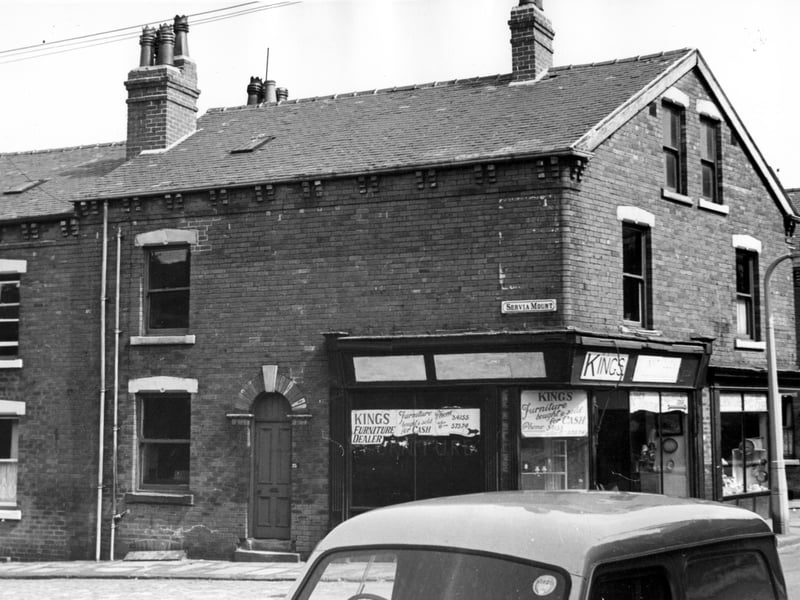 To the left is number 21 Servia Mount with 23 next to the shop. This house appears to be empty. Kings furniture shop is number 15 Servia Road. Sign in the window reads 'furniture bought and sold for cash'. The adjacent window of number 17 can be seen at the junction with Servia Place.