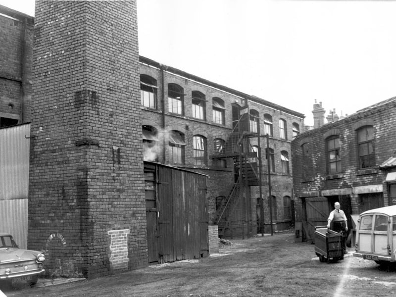 Roscoe Clothing Clothing Co Ltd factory, the front was at Meanwood Road. Access to this yard was from Oxford Road. The base of the factory chimney is on the left. This photo was taken in August 1967, on what must have been a warm day, all the windows are open and emergency doors to the fire escape are propped open. On the right is a workman with a trolley.
