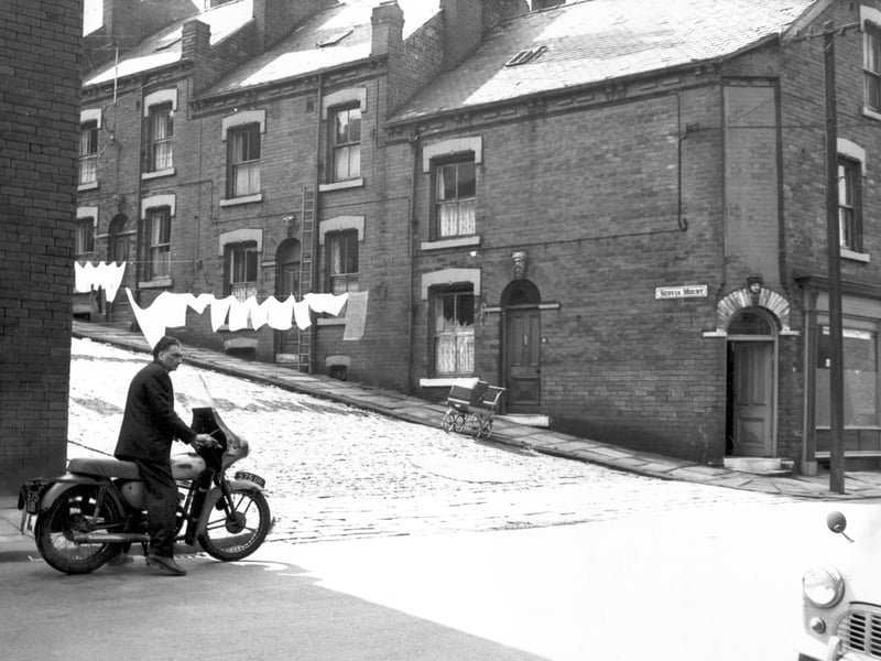 Servia Mount on the left is number 30, moving right are 28 then 26 at the end of the row. The premises on the corner at the junction with Servia Road also numbered 26. On the left is a man with a motor bike.
