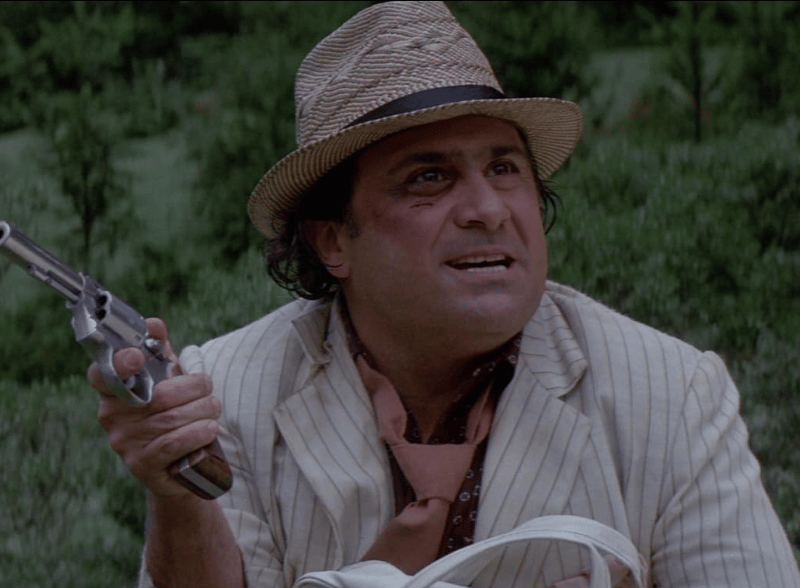 DeVito’s character, Ralph, is a small-time thief who complicates the lives of the main characters, played by Michael Douglas and Kathleen Turner. His bumbling antics and greedy demeanour provide comic relief, offering a stark contrast to the romantic adventure unfolding in the jungle. His role was integral to the film’s success as a fast-paced action-comedy.
