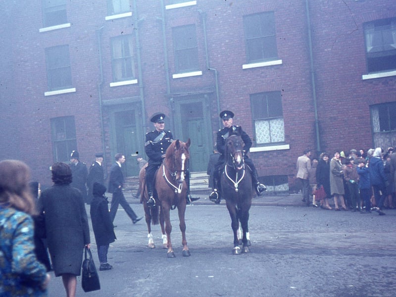 Two policemen on horseback with other police in the background. Groups of people are standing nearby in anticipation of Queen Elizabeth II visit to Leeds to open Seacroft Civic Centre. The houses pictured are on Portland Crescent and have since been demolished. Pictured in October 1965.