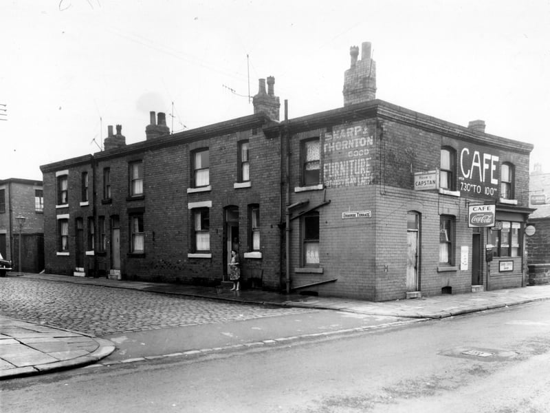 Ormonde Terrace and Sackville Street in October 1960. An electricity sub-station is situated at the left edge, then three houses on Ormonde Terrace. A woman is outside with a bucket, the pavement has just been cleaned with water. This was called 'swilling'. The cafe is 13/15 Sackville Street, a painted sign on the wall to Ormonde Terrace is for Sharp and Thorton furniture, the store was on Vicar Lane in Leeds. To the right is Sheepscar Beck with steps leading down to footpath.