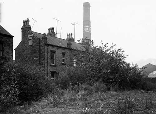 Rear view of numbers 9 to 13 Royds Lane. Wortley Beck runs on the right side of this scene, past the houses, out of view on the right is a reservoir adjacent to the beck. A large mill chimney with the name Fox painted on can be seen.