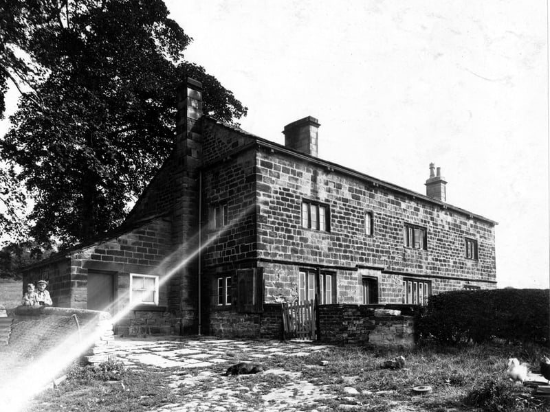The front view of the farmhouse of Pigeon Cote Farm in August 1948. The building is stone-built and has a flagged yard, where a dog lies asleep. On the left, a man and a child look over a stone wall at the camera. A chicken is on the right.