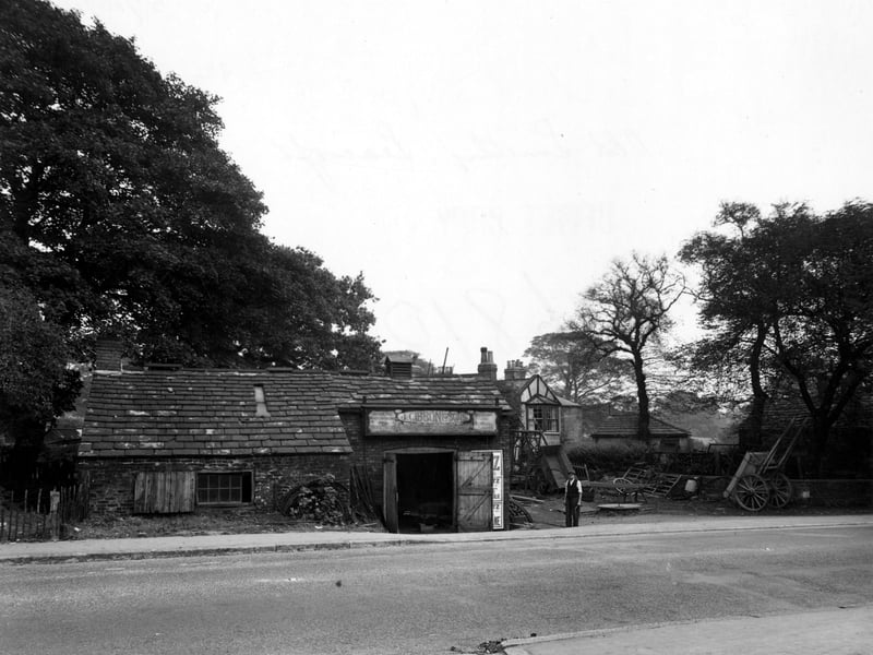 Old Smithy on N.E side of York Road, handcarts in yard to right of building, a man is standing outside. Just behind the Smithy is a half-timbered building with large trees on either side. Pictured in October 1949.