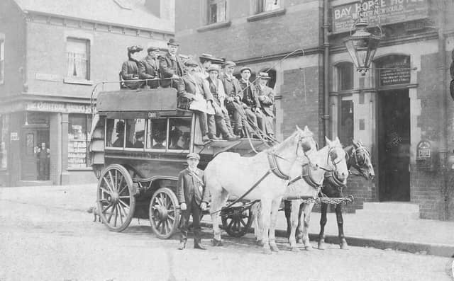 A horse-drawn omnibus stands outside the Bay Horse Hotel on Benson Place in June 1901. The omnibus, proprietor, John Thomas Bean & Son, is loaded with passengers on both the upper and lower deck. It is drawn by three horses. Mr. Bean, seated in the centre with whip, is dressed identically to the young man standing, most likely his son. They are wearing suits, cloth caps and are sporting button holes.