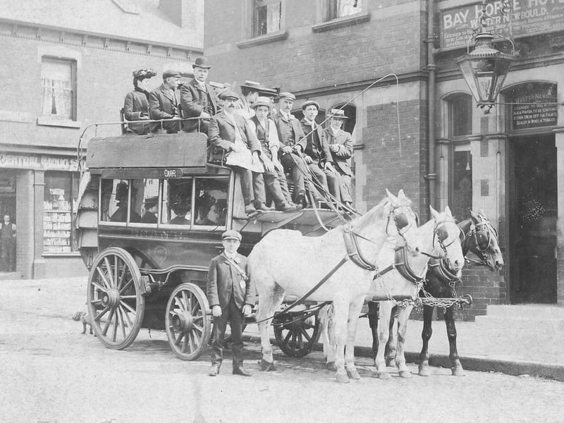 A horse-drawn omnibus stands outside the Bay Horse Hotel on Benson Place in June 1901. The omnibus, proprietor, John Thomas Bean & Son, is loaded with passengers on both the upper and lower deck. It is drawn by three horses. Mr. Bean, seated in the centre with whip, is dressed identically to the young man standing, most likely his son. They are wearing suits, cloth caps and are sporting button holes.