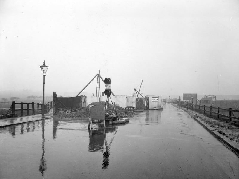 The construction of a railway bridge over Wortley Beck on Gelderd Road. The photo from October 1935 was taken at 11.30am showing large mound of dug earth in the road. A two light traffic light is stopping traffic. A car is coming from the left. On the right is a fenced off field. In the background on the right is a 'No Footpath', sign and an 'Evening Post' billboard.