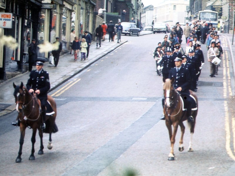 The procession for the last annual Mayor's Sunday Service parade before the abolition of Morley Town Council makes its way down Queen Street to St. Peter's Parish Church. Policemen on horses lead the way, followed by a group of marching policemen then scouts and guides. Shops seen on the left include Wing Hong Chinese restaurant.