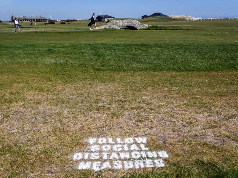 A golfer crosses the iconic Swilcan Bridge with a sign on the grass reminding golfers to respect social distancing measures at The Old Course in St Andrews.