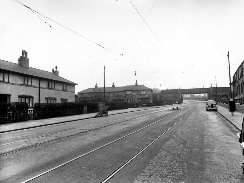 Belle Isle Road in November 1950. Looking south towards an iron railway bridge. On the right of the road there is a brick industrial building with a wall and iron fence running along the pavement. There is a woman walking along the pavement and a car parked at the kerb.